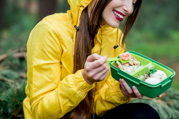 Junge Frau, die während des Reisens im Wald einen Snack mit gesundem Essen in der Lunchbox isst
