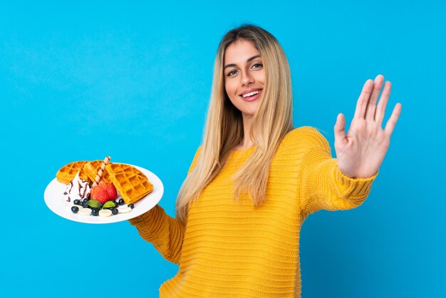 Junge Frau, die Waffeln über isolierter Wand hält, die mit Hand mit glücklichem Ausdruck salutiert