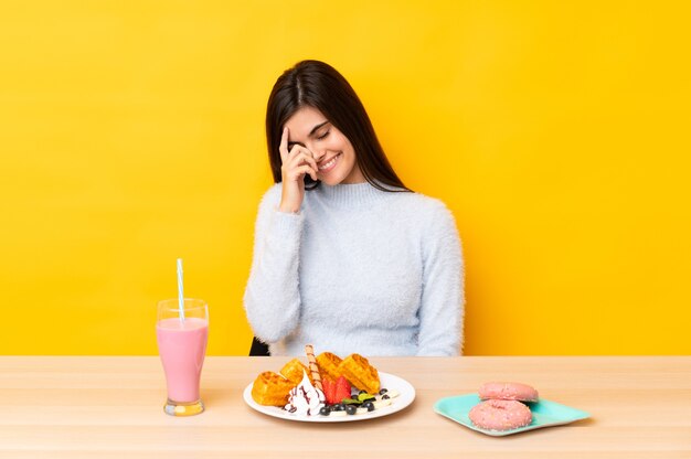 Junge Frau, die Waffeln und Milchshake in einem Tisch über isoliertem gelbem Lachen isst