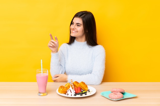 Junge Frau, die Waffeln und Milchshake in einem Tisch über isoliertem gelbem Zeigefinger zur Seite isst