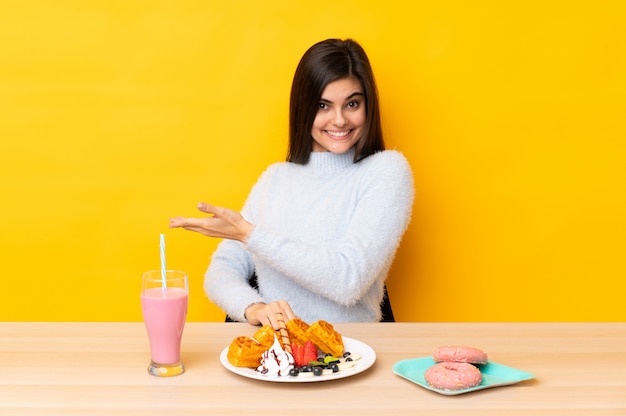 Junge Frau, die Waffeln und Milchshake in einer Tabelle über Ausdehnungshänden der lokalisierten gelben Wand zur Seite für die Einladung isst zu kommen