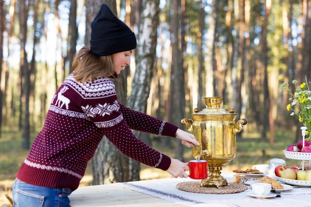 Junge Frau, die warmes Trikot trägt, das etwas Tee mit Weinlesesamowar am Wald gießt
