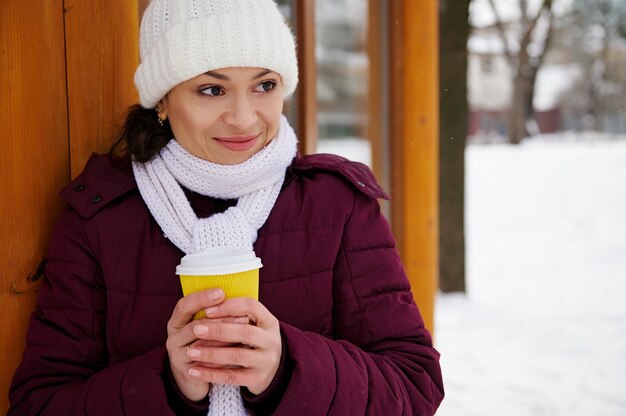 Junge frau, die weißen schal und weißen wollhut trägt, der mit einem heißen getränk steht, um an einem winter-schneepark zu gehen.