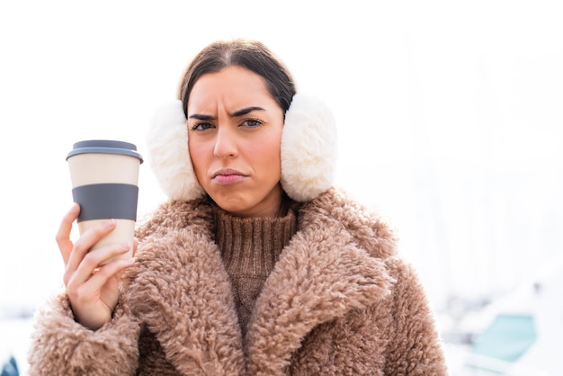 Junge Frau, die Wintermuffs trägt und mit traurigem Gesichtsausdruck Kaffee zum Mitnehmen im Freien hält