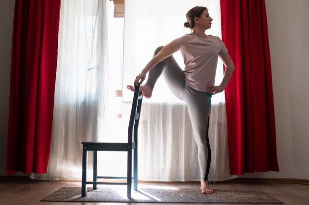 Junge Frau, die Yoga macht und dabei ein einsames Bein mit einem Stuhl balanciert