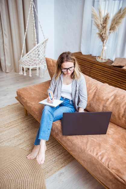 Junge Frau, die zu Hause auf Sofa sitzt. Positives Mädchen schreibt in Notebook und tippt auf Notebook-Tastatur. Online-Ausbildung, Ausbildung und freiberufliche Tätigkeit. Computer, Laptop und Fernstudium