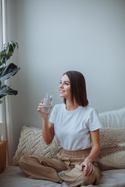 Junge Frau, die zu Hause mit einem Glas Wasser auf dem Bett sitzt