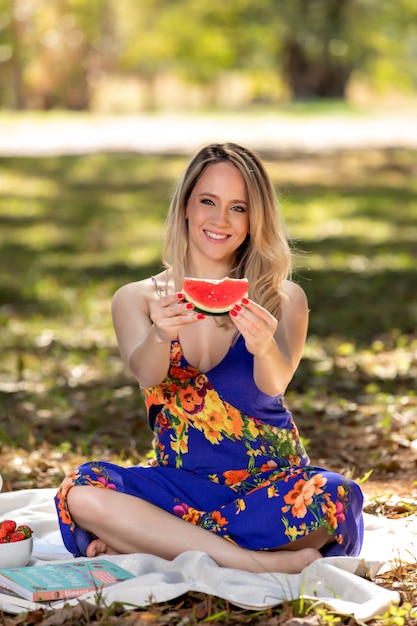 Junge Frau draußen unter Bäumen in einem Park, glücklich und lächelnd, Picknick machend und Wassermelone schmeckend.