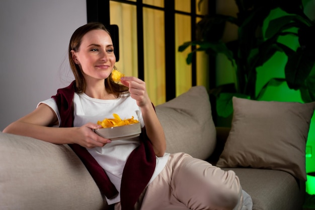 Junge Frau entspannt sich auf der Couch, sieht fern und isst Chips