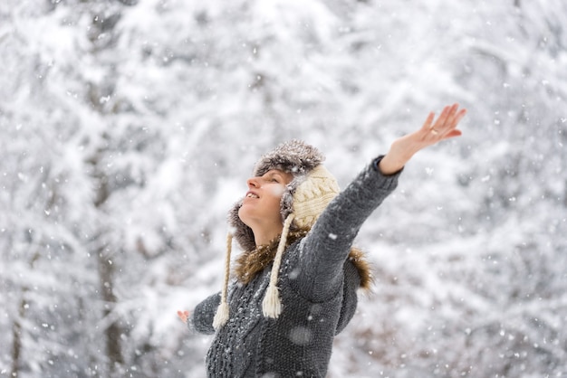 Junge Frau feiert den Winter im Freien in einem winterlichen Garten im fallenden Schnee mit ausgebreiteten Armen.