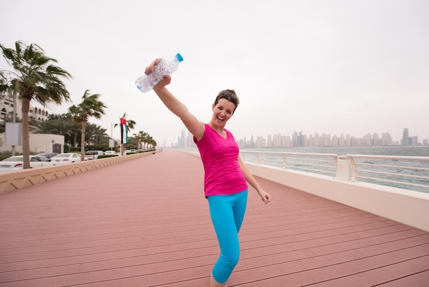 Junge Frau feiert einen erfolgreichen Trainingslauf auf der Promenade am Meer mit einer Flasche Wasser und erhobenen Händen mit einer Großstadt im Hintergrund