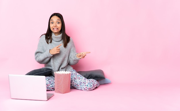 Junge Frau gemischter Abstammung, die Popcorn isst, während sie einen Film auf dem Laptop erschrocken ansieht und zur Seite zeigt