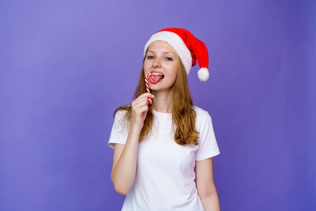 Junge Frau genießt Süßigkeiten in Weihnachtsmütze und weißem T-Shirt auf lila Hintergrund Mädchen kaut auf einem Chris...