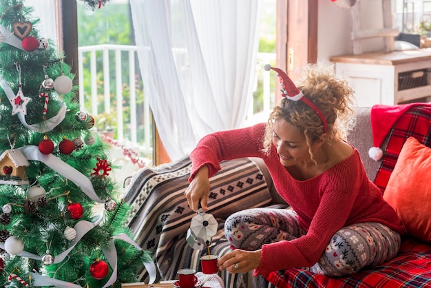 Junge Frau gießt Kaffee aus dem Wasserkocher in eine Tasse, während sie zu Hause auf dem Sofa im Wohnzimmer frühstückt. Kaukasische Frau, die zu Hause Weihnachten mit verziertem Baum feiert.