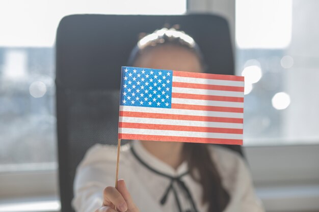 Junge Frau hält amerikanische Flagge in der Hand, sitzt in einem Sessel in einem Büro an einem Tisch