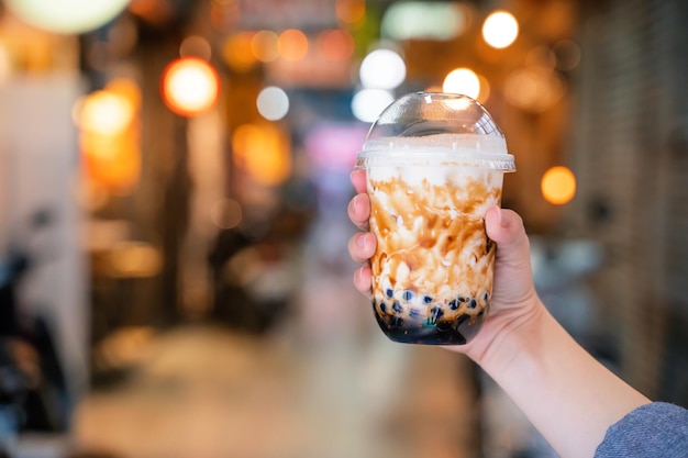 Junge Frau hält das Trinken von Tapioka-Perlblasen-Milchtee mit braunem Zuckergeschmack mit Glasstroh im Nachtmarkt von Taiwan in der Nähe von Bokeh