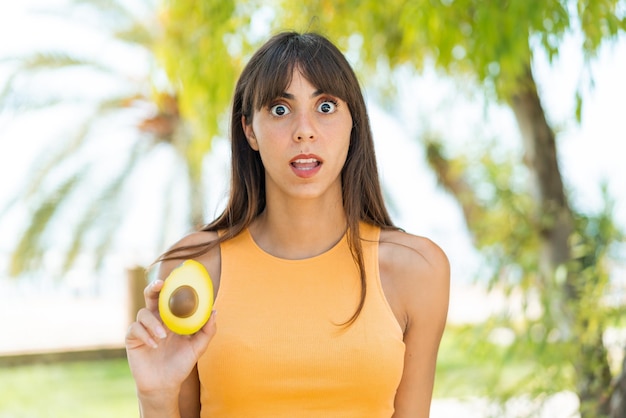 Junge Frau hält eine Avocado im Freien mit überraschtem und schockiertem Gesichtsausdruck