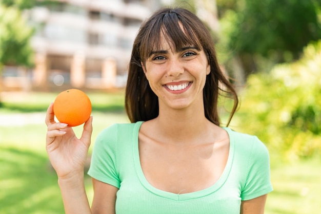 Junge Frau hält eine Orange im Freien und lächelt viel