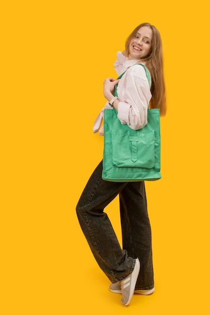Junge Frau hält grüne, umweltfreundliche Einkaufstasche in ihrer Schulter, gelber Hintergrund, vertikaler Rahmen