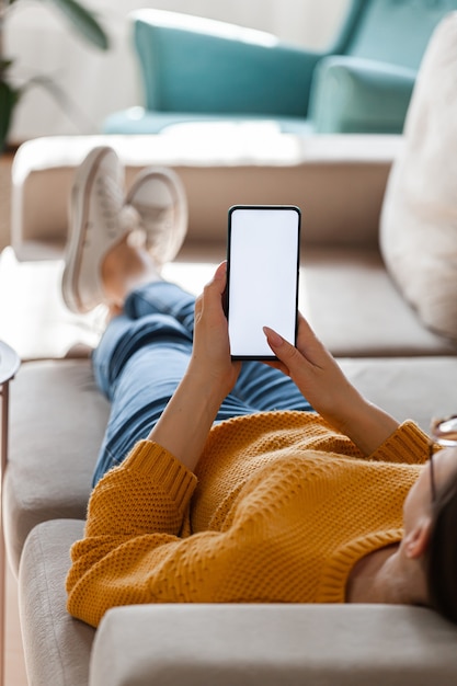 Junge Frau hält Handy in Händen, auf dem Sofa liegend