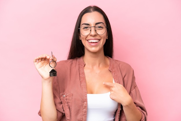 Junge Frau hält Hausschlüssel isoliert auf rosa Hintergrund und überraschtem Gesichtsausdruck
