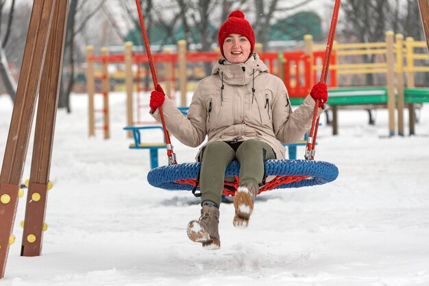 Junge Frau hat Spaß im Winter im Freien Süßes Mädchen in roter Strickmütze und Fäustlingen reitet fröhlich auf dem Schaukelspielplatz