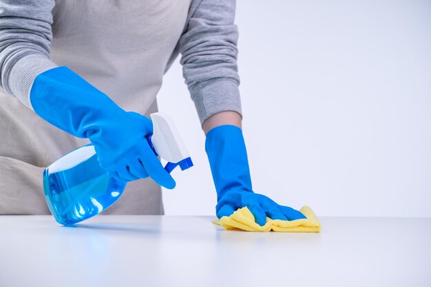 Junge Frau Haushälterin reinigt weißen Tisch in Schürze mit blauen Handschuhen Sprühreiniger nassen gelben Lappen Nahaufnahme Kopierraum leeres Designkonzept