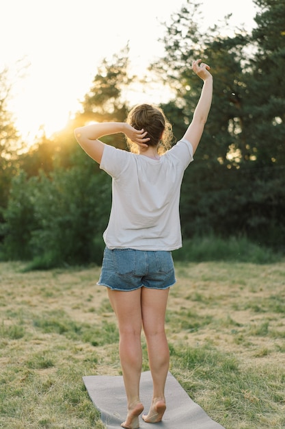 Junge Frau hebt Ihre Hände bei sonnigem Wetter auf der Wiese bei Sonnenuntergang im Freien im Sommer entspannen