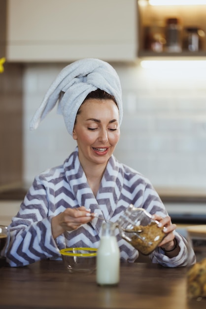 Junge Frau im Bademantel bereitet in der heimischen Küche ein Müsli zum Frühstück zu.