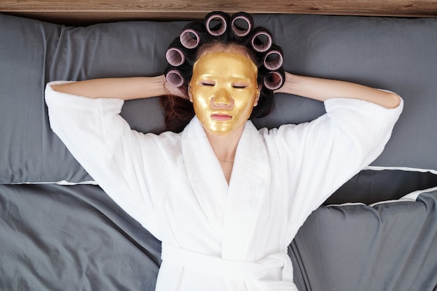 Junge Frau im Bademantel, die sich auf dem Bett mit Haarrollen und Lakenmaske entspannt, Blick von oben