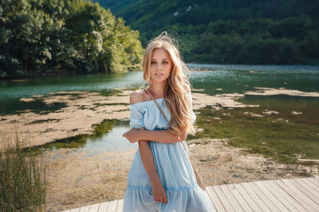 Foto junge frau im blauen kleid, das allein auf fußgängerbrücke steht und auf see starrt nebel über wasser kühler morgen schönes modisches mädchen mit einer strohtasche, die auf einem bergsee posiert