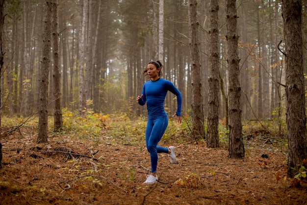 Junge Frau im blauen Trainingsanzug, die im Herbst auf dem Waldweg in Richtung Kamera läuft