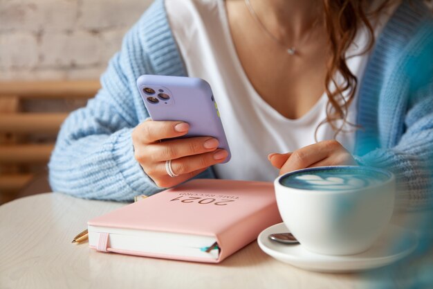 Junge Frau im blauen warmen Pullover, der eine Textnachricht vom Handy beim Kaffeetrinken liest. Geschäftsfrau überprüft soziale Medien auf ihrem Smartphone. Blogger-Lebensstil. Planen und organisieren