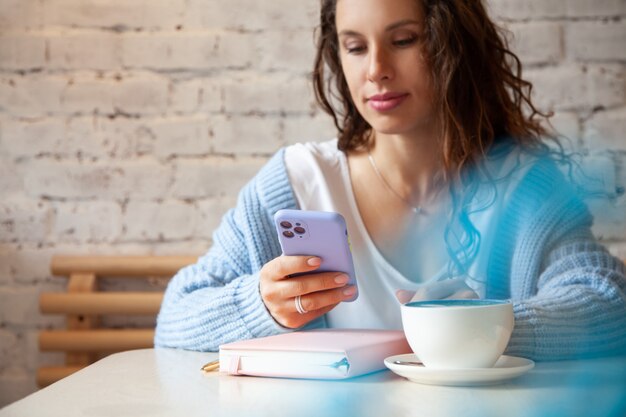 Junge Frau im blauen warmen Pullover, der eine Textnachricht vom Handy beim Kaffeetrinken liest. Geschäftsfrau überprüft soziale Medien auf ihrem Smartphone. Blogger-Lebensstil. Planen und organisieren