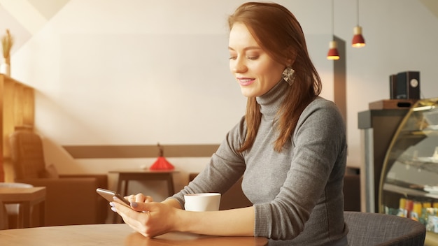 Junge Frau im Café tippt eine Nachricht am Telefon und trinkt eine Tasse Kaffee, die auf jemanden wartet. Seitenansicht. Sonnenlicht
