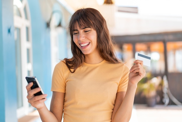 Junge Frau im Freien kauft mit dem Handy und einer Kreditkarte ein