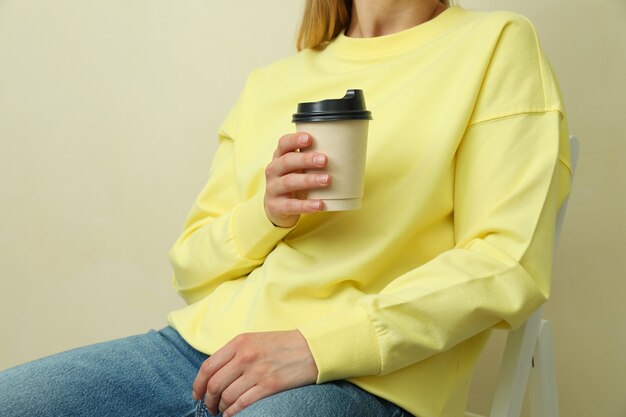 Junge Frau im gelben Sweatshirt, das mit Pappbecher gegen beige Oberfläche sitzt