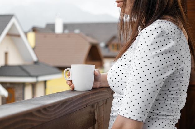 Junge Frau im gepunkteten T-Shirt mit einer weißen Tasse Tee oder Kaffee, die auf dem Balkon steht.