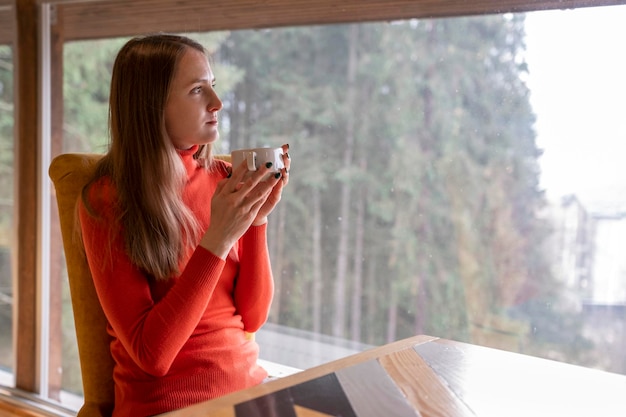 Junge Frau im hellen Rollkragenpullover entspannt sich im Café und trinkt Tee Besucher im Restaurant sitzt am Fenster mit Blick auf den Wald und hält Tasse