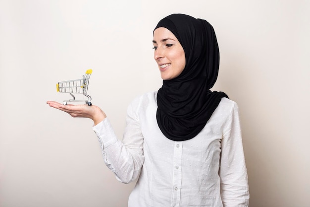 Junge Frau im Hijab mit einem Einkaufswagen auf hellem Hintergrund