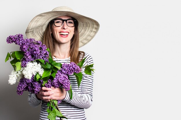 Junge Frau im Hut und in den Gläsern glücklich hält einen Blumenstrauß auf einem hellen Hintergrund.