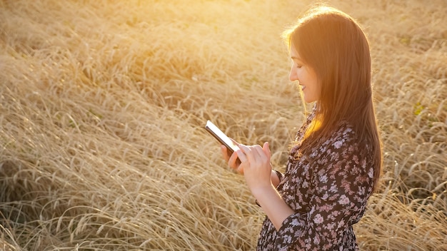 Junge Frau im Kleid schaut auf das Telefon, während sie in einem Weizenfeld-Exemplar steht