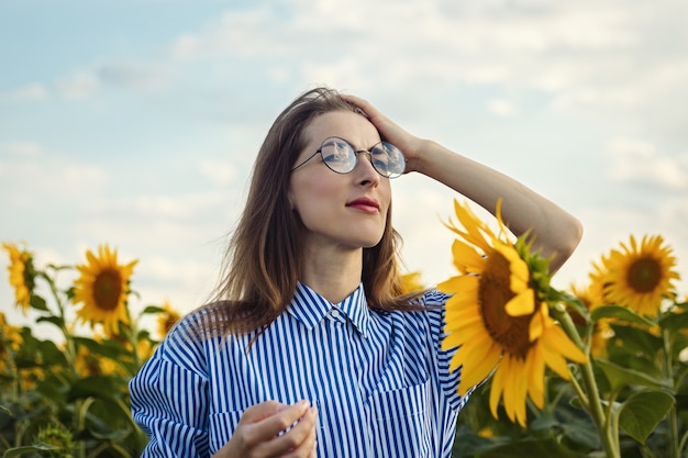 Junge Frau im Kleid und in den Gläsern, die den Sonnenuntergang auf dem Sonnenblumenfeld genießen.