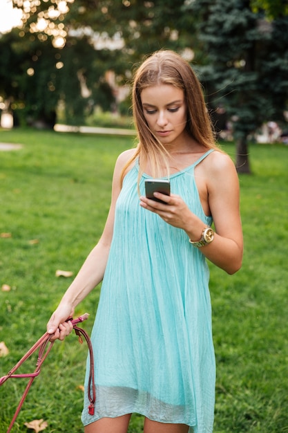 Junge Frau im Kleid unter Verwendung des Mobiltelefons beim Gehen ihres Hundes im Park