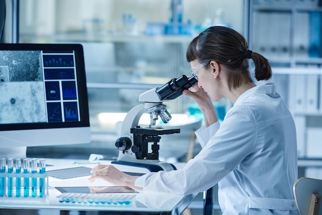 Junge Frau im Laborkittel, die während ihrer Arbeit im Labor durch das Mikroskop schaut, während sie mit Computer und Reagenzgläsern am Tisch sitzt