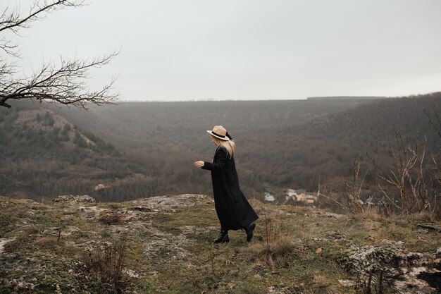 Junge Frau im langen schwarzen Mantel und im beige Hut im Freien, die auf dem Hintergrund der Berge gehen.