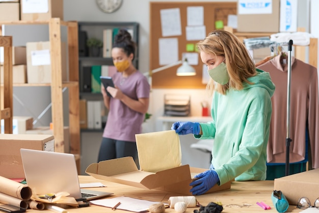 Foto junge frau im maskenpackpaket während beim stehen in der nähe des tisches mit ihrem kollegen in der wand in der werkstatt