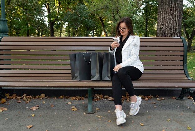 Junge Frau im Park mit Einkaufstüten und Telefon. Online Einkaufen. Rabatte, Aktionen, schwarzer Freitag.