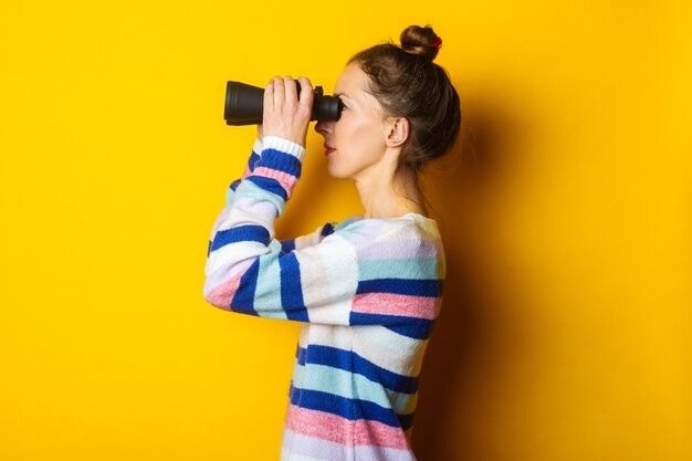 Junge Frau im Pullover, der durch Fernglas auf gelbem Hintergrund schaut.