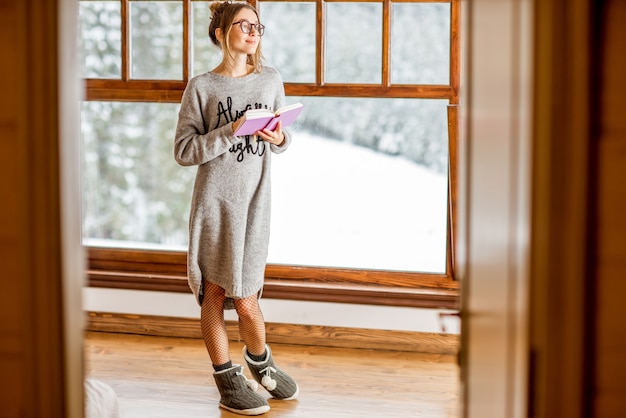 Junge Frau im Pullover, die in der Nähe des großen Fensters im gemütlichen Berghaus aus Holz mit schönem Landschaftsblick während der Winterzeit steht